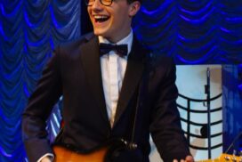 AJ as Buddy, in black suit with bow tie, black glasses, guitar, smiling widely.