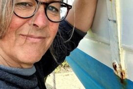 Close up of Nic, wearing glasses, smiling and leaning against the hull of a boat.